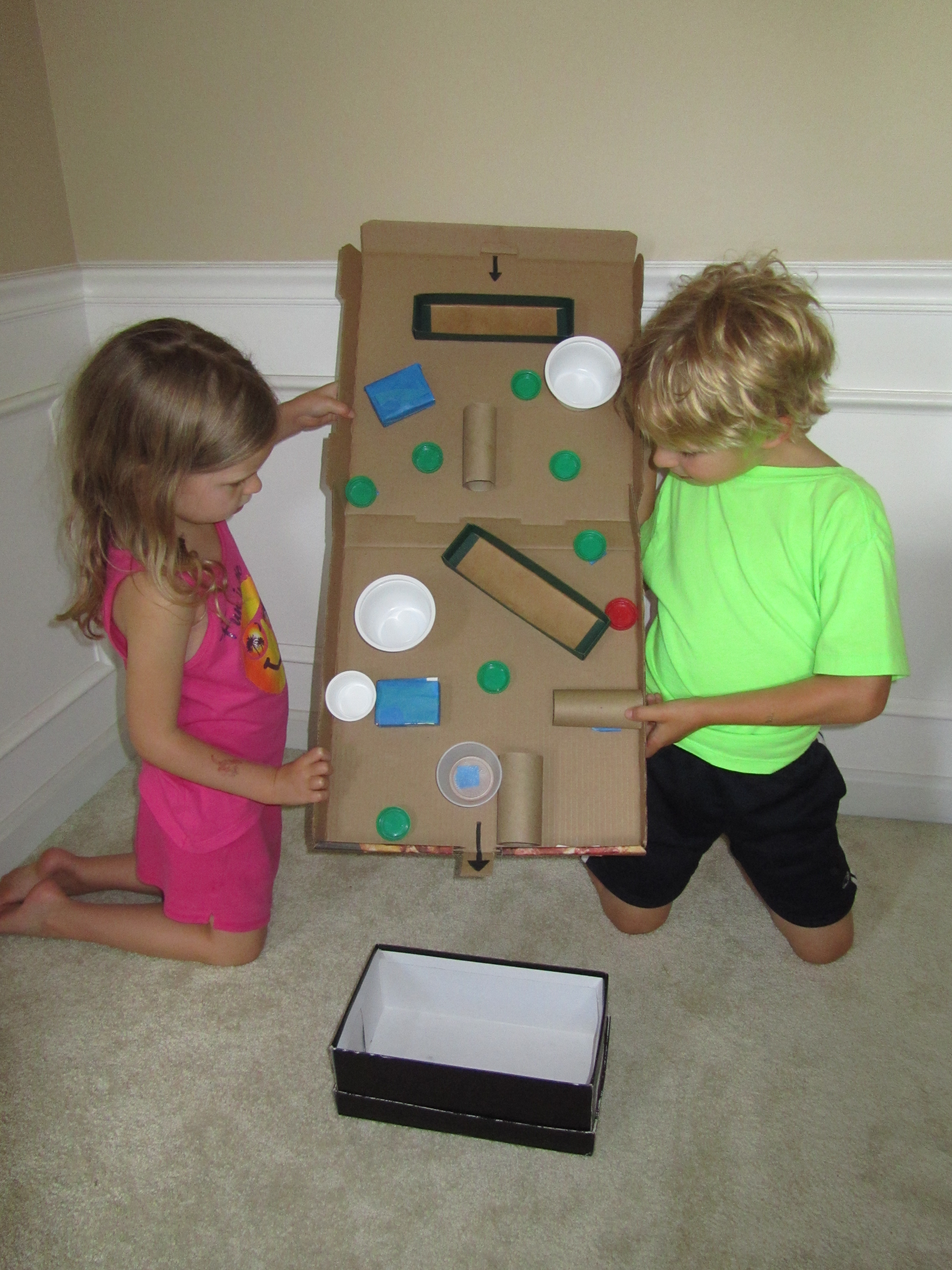 Two children work coopertively to move the red cap through a maze created from recycled plastic and cardboard materials